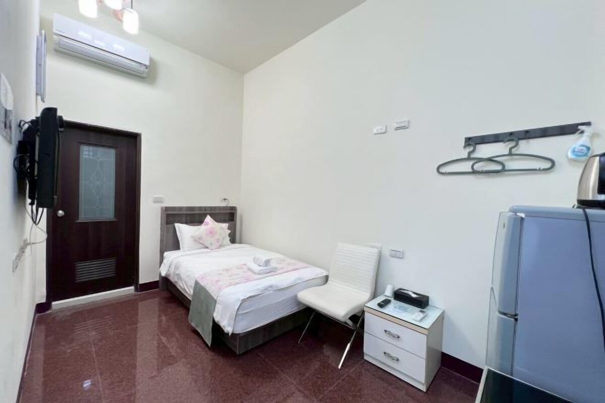 A hotel room with a bed, bedside table, a small desk, a chair, a TV, and a refrigerator.