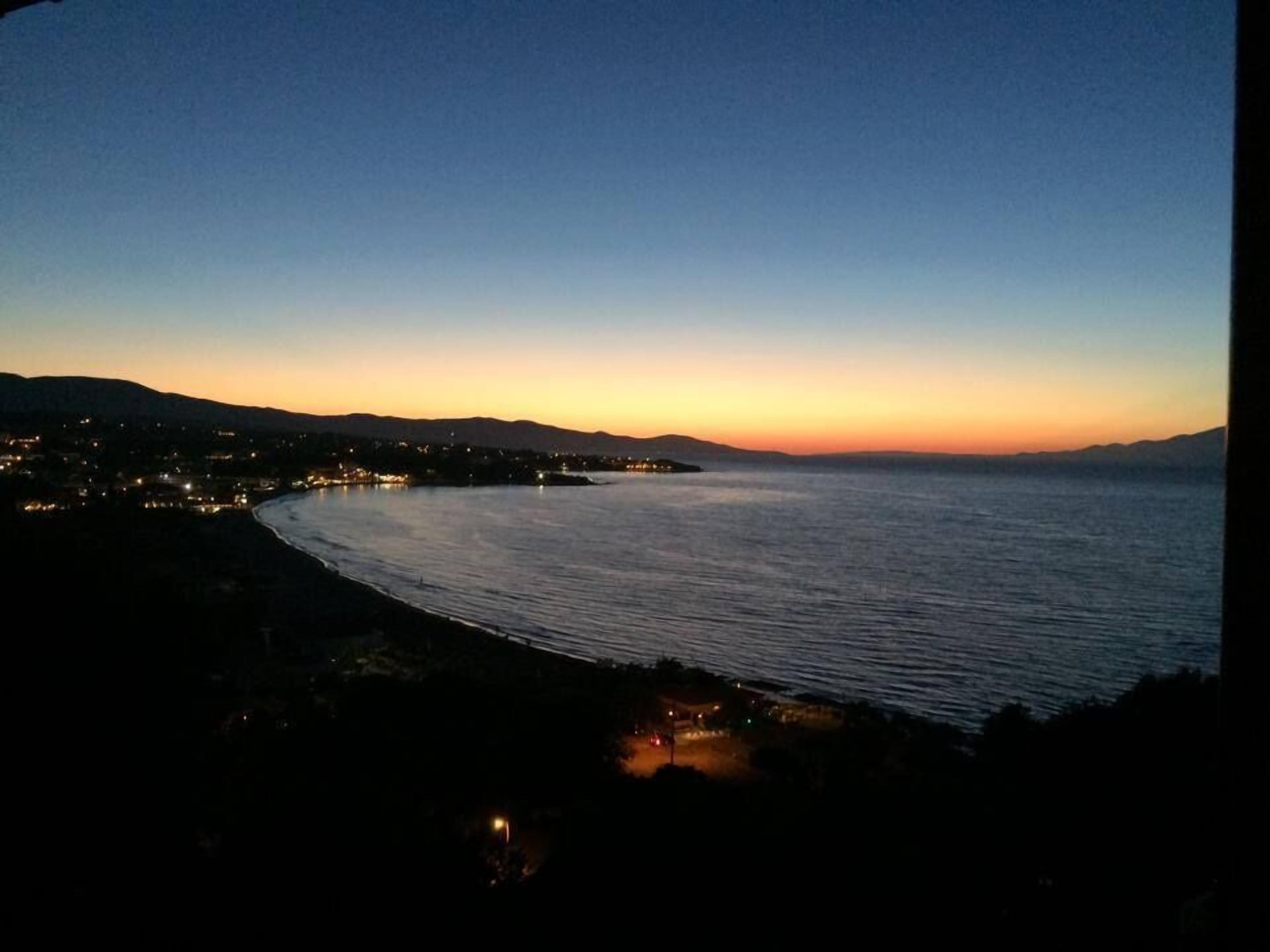 A coastal cityscape at dusk with a calm sea, distant hills, and a few illuminated buildings along the shoreline.