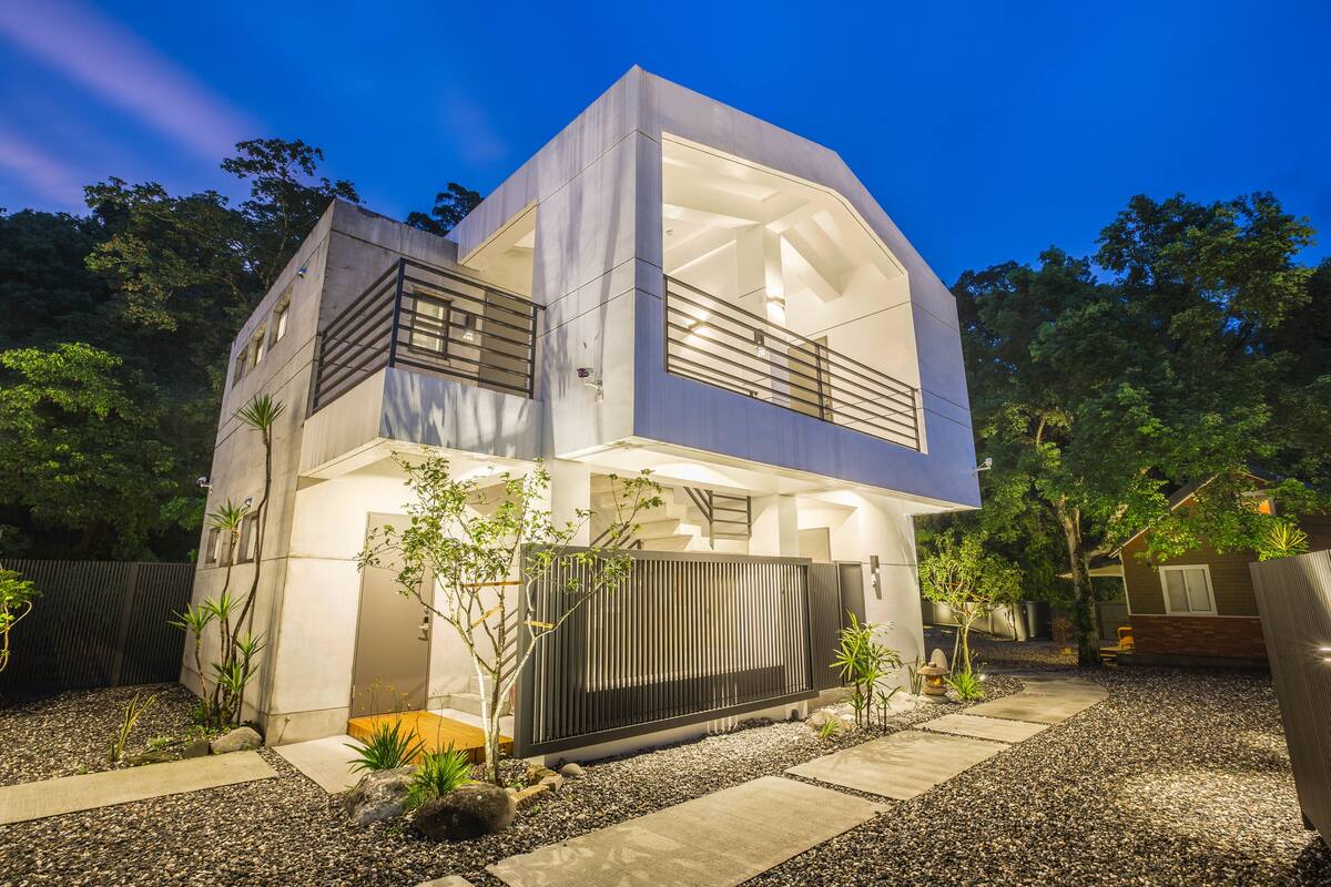 Uma casa moderna de dois andares com varanda, cercada por um caminho de cascalho e pequenas plantas.