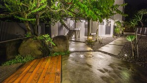Un espacio al aire libre de noche con una terraza de madera, senderos de concreto y un muro de piedra.