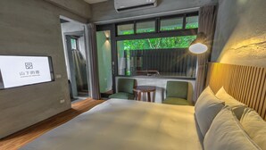 Una habitación de hotel con una cama grande, una mesita pequeña y un sofá. Cuenta con una ventana con cortinas, un televisor montado en la pared y un ventilador de techo.