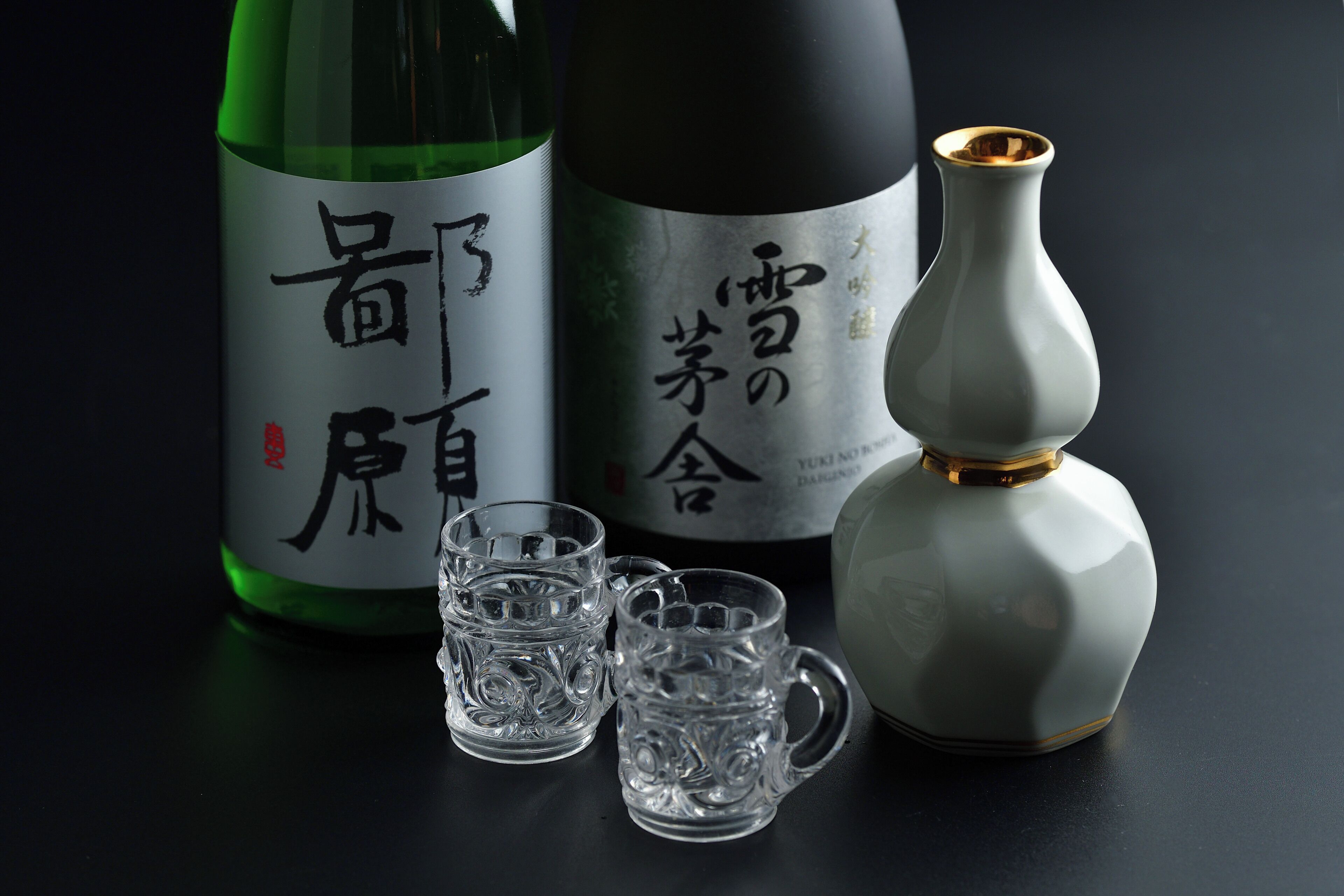 A bottle of sake, two small glasses, and a decorative vase on a dark surface.