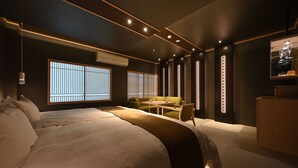 Habitación de hotel moderna con una cama grande, un sofá, una mesita y un escritorio. Se observan ventanas con persianas verticales, un aire acondicionado instalado en la pared y un estante con objetos decorativos.