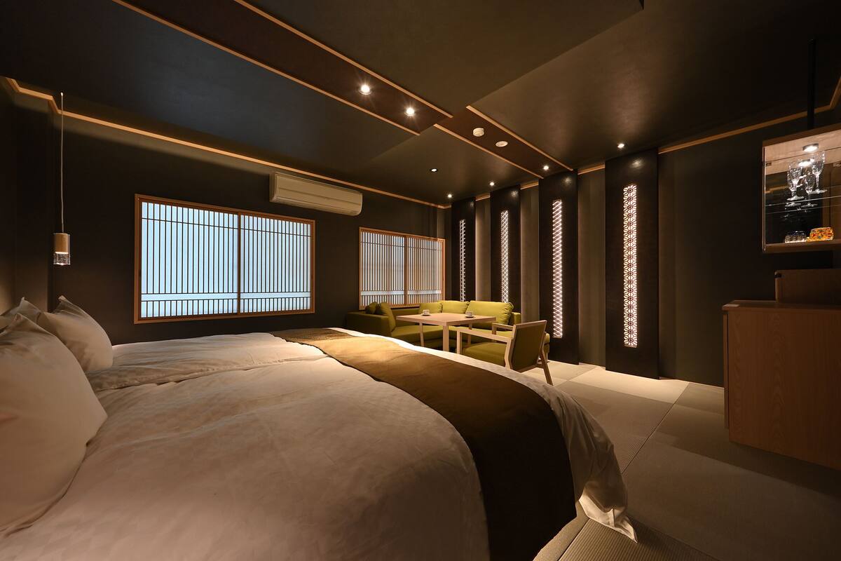 A modern hotel room with a large bed, a sofa, a small table, and a desk. There are windows with vertical blinds, a wall-mounted air conditioner, and a shelf with decorative items.