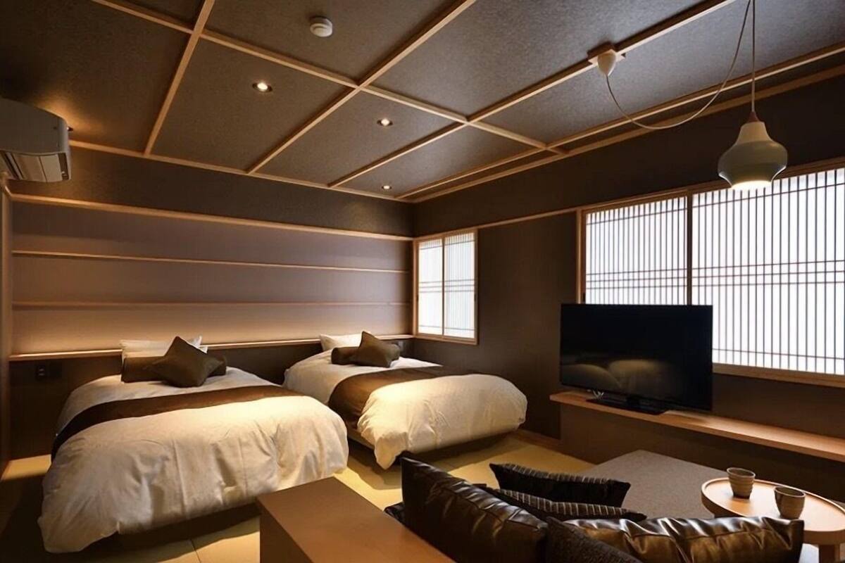 A modern hotel room with two beds, a flat-screen TV, a sofa, and a large window with blinds.