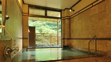 A traditional Japanese onsen with a wooden interior, a view of a forested area through the sliding door, and a stone floor.