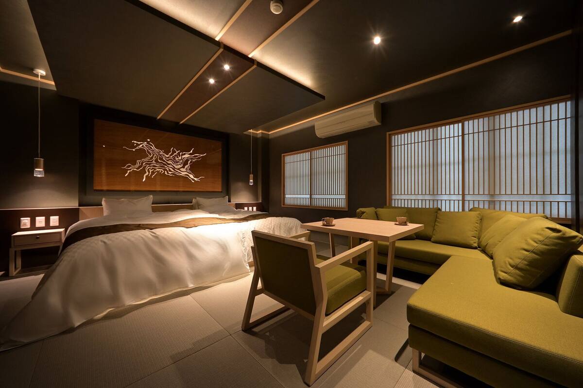 A modern hotel room with a large bed, a green sofa, a small table, and a wall-mounted artwork.