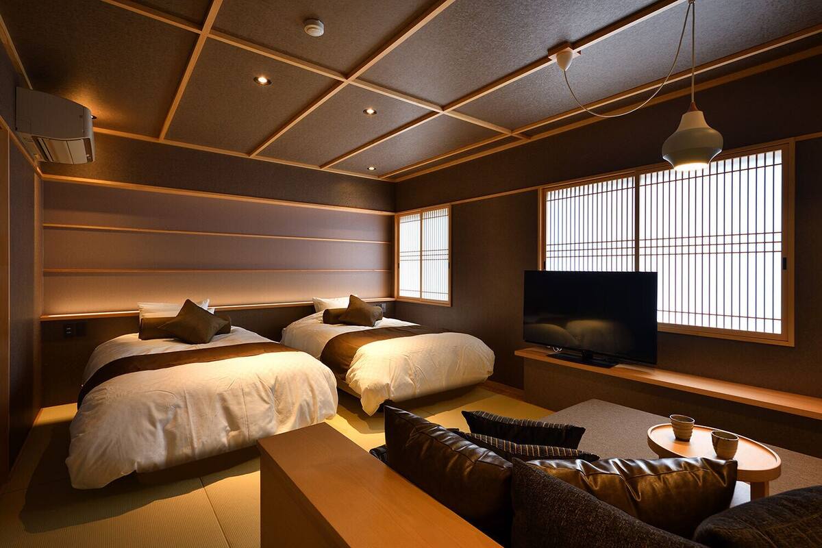 A modern hotel room with a large bed, a sofa, a TV, and a window with blinds.