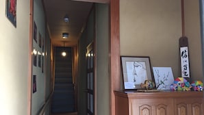 A hallway with a staircase leading to a landing. There are framed artworks on the walls, a wooden cabinet with decorative items, and a sign with Chinese characters. - Kyoto Guesthouse Oyado KEI (Kyoto)