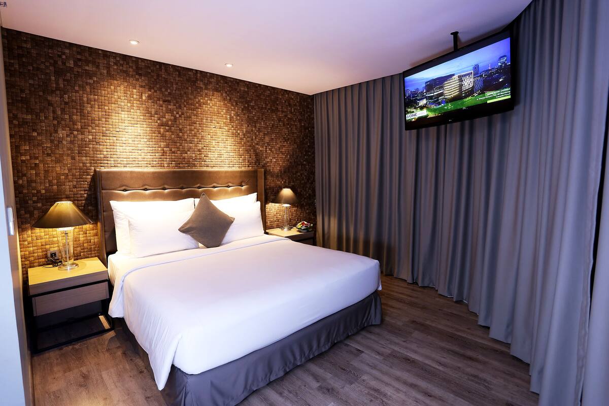 A modern hotel room with a brick-patterned wall, a large bed with white linens, two bedside lamps, a flat-screen TV mounted on the wall, and a wooden floor.