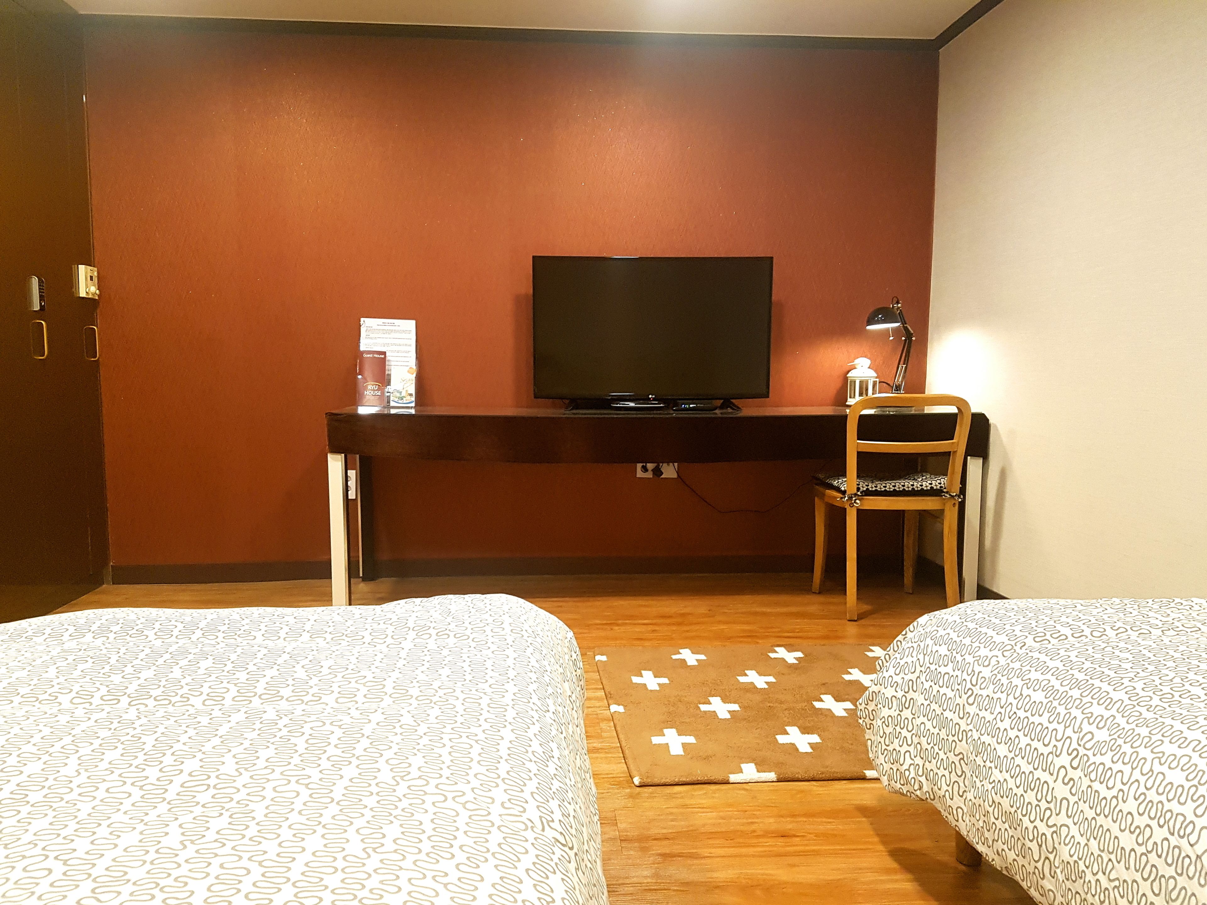 A hotel room with two beds, a desk with a TV, a chair, and a small table with a lamp.