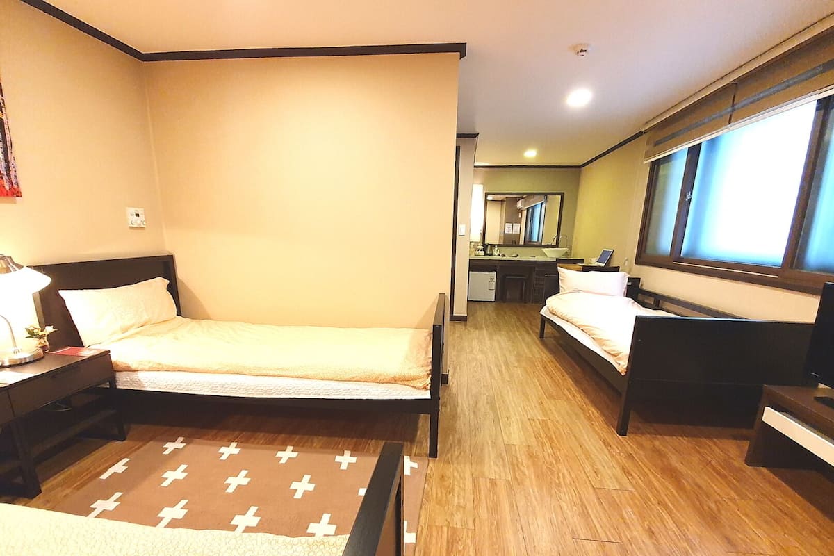 A hotel room with two single beds, a desk with a lamp, a small kitchenette, and a large window.