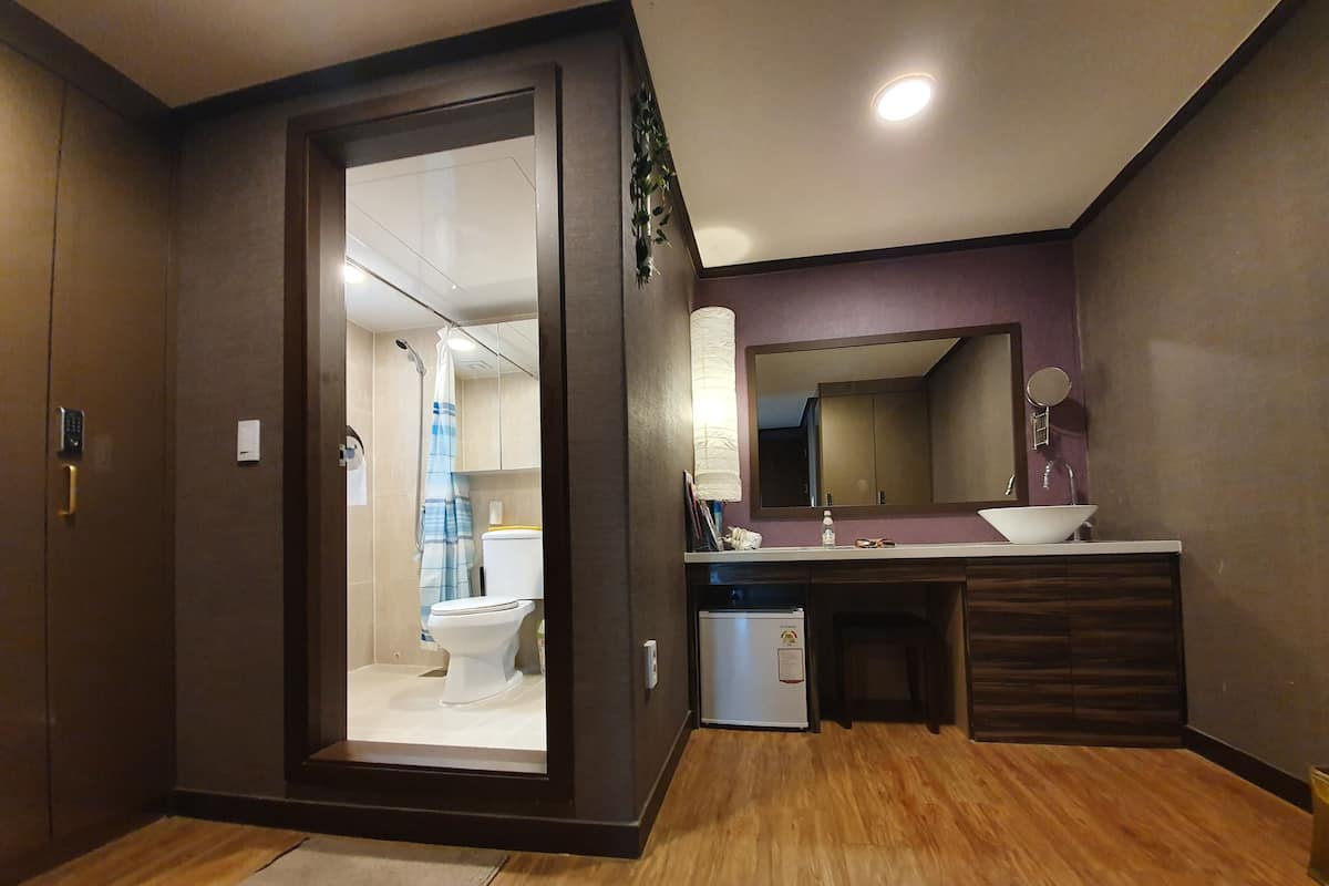 A modern bathroom with a wooden floor, a white sink, a mirror, and a toilet visible through a glass door.