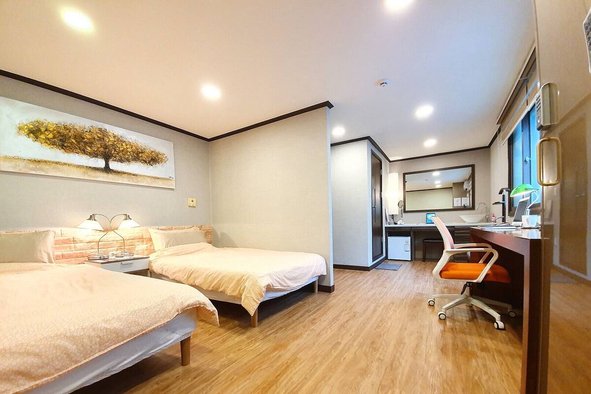 A hotel room with two beds, a desk with a chair, a small kitchenette, and a large window.
