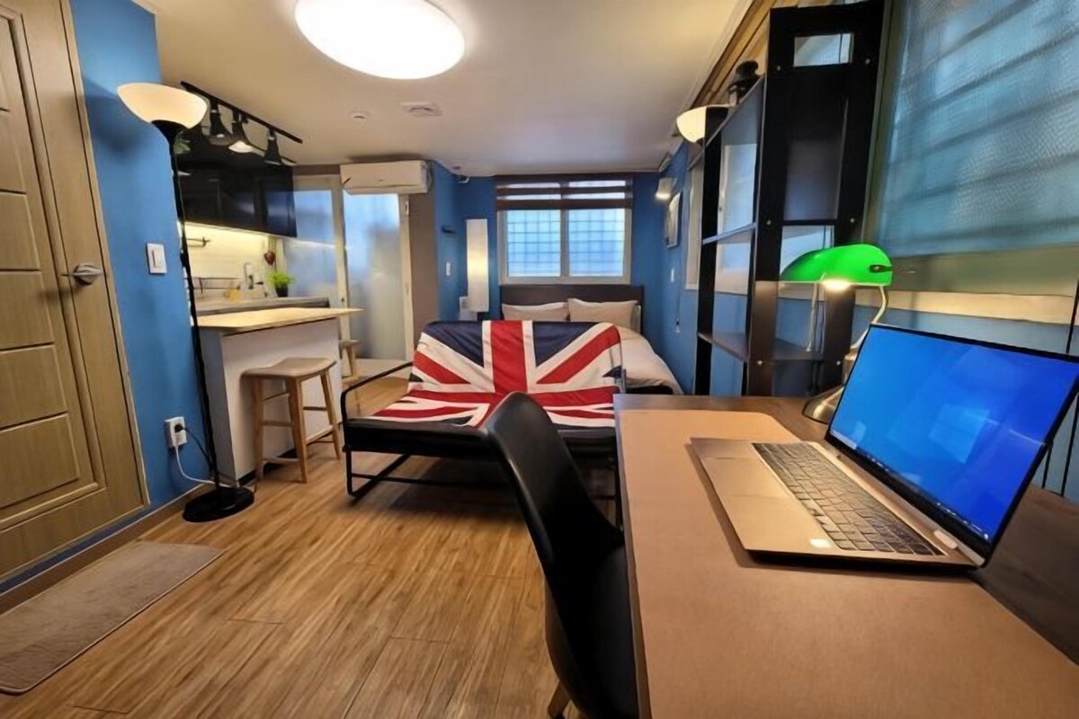 A compact room with a bed, a desk with a laptop, a chair, a small stool, and a green desk lamp.