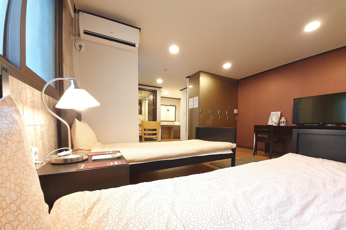 A hotel room with a bed, bedside lamp, desk, chair, TV, wardrobe, and an air conditioning unit.