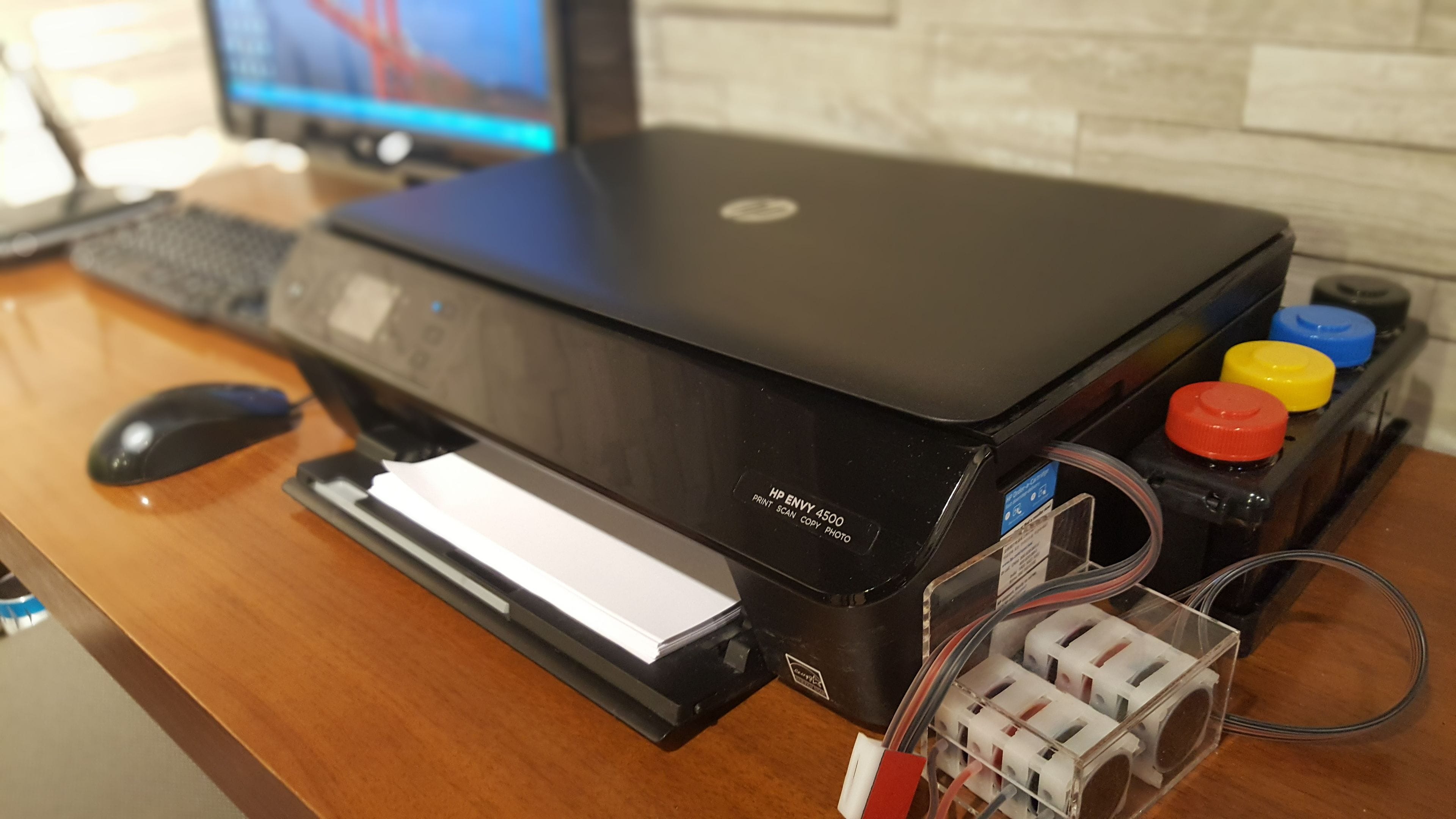 Una impresora HP ENVY 4500 negra sobre un escritorio de madera, con un ratón, un teclado y un monitor al fondo.