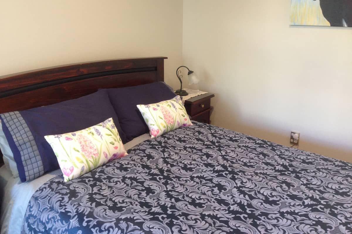 A neatly made bed with a patterned bedspread and two pillows, a wooden headboard, a bedside table with a lamp, and a wall-mounted painting of a horse and bird.