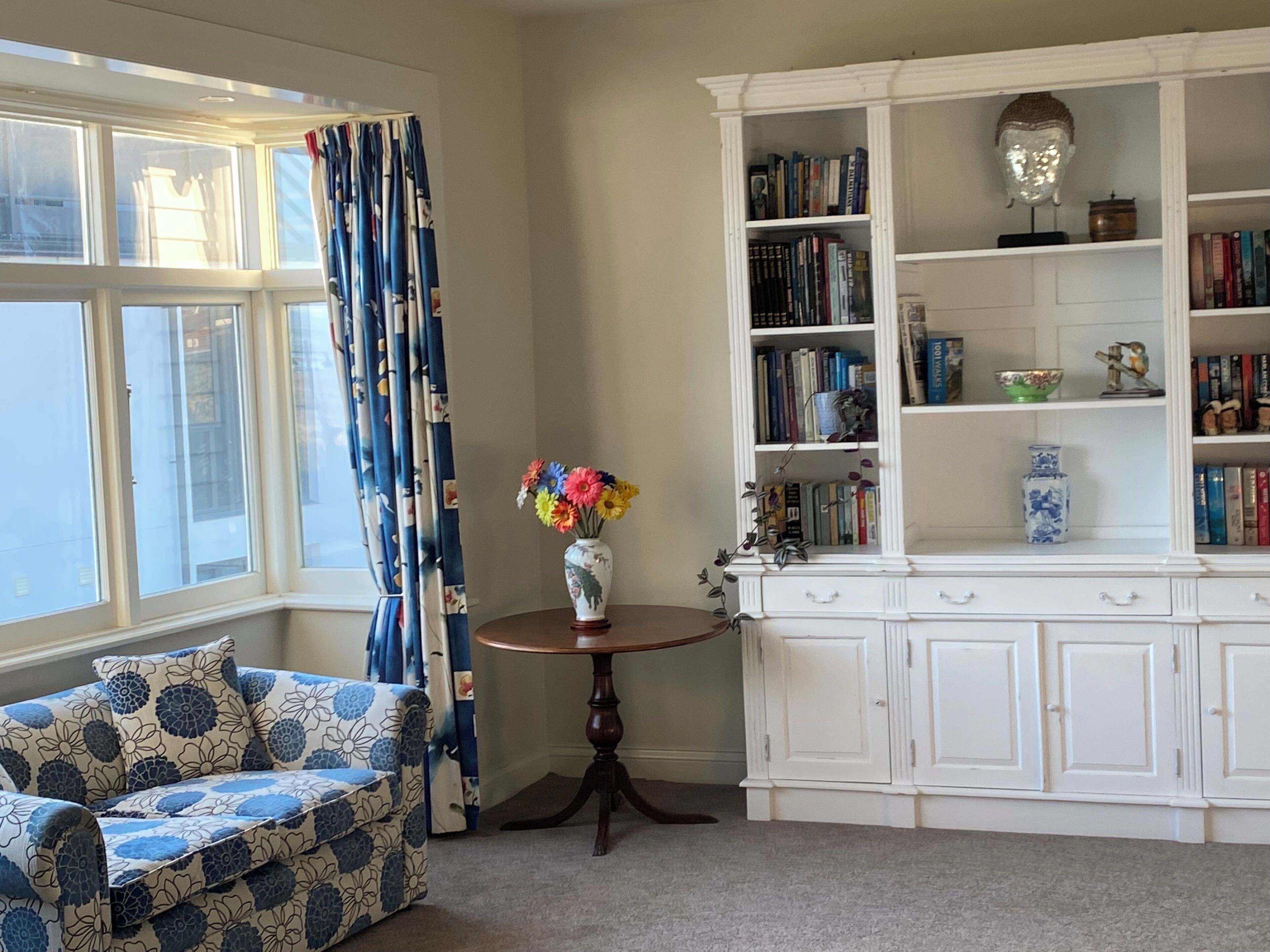 A room with a patterned sofa, a small round table with a vase of flowers, a white bookcase, and a window with curtains.