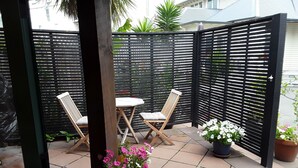 Uma área de pátio com duas cadeiras de madeira e uma pequena mesa, cercada por um muro preto e plantas em vasos.