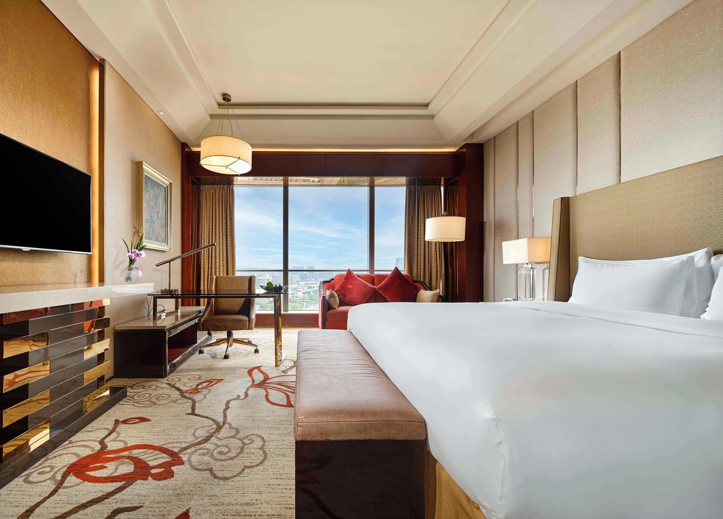 A hotel room with a large bed, a desk, a chair, a TV, and a view of the city.