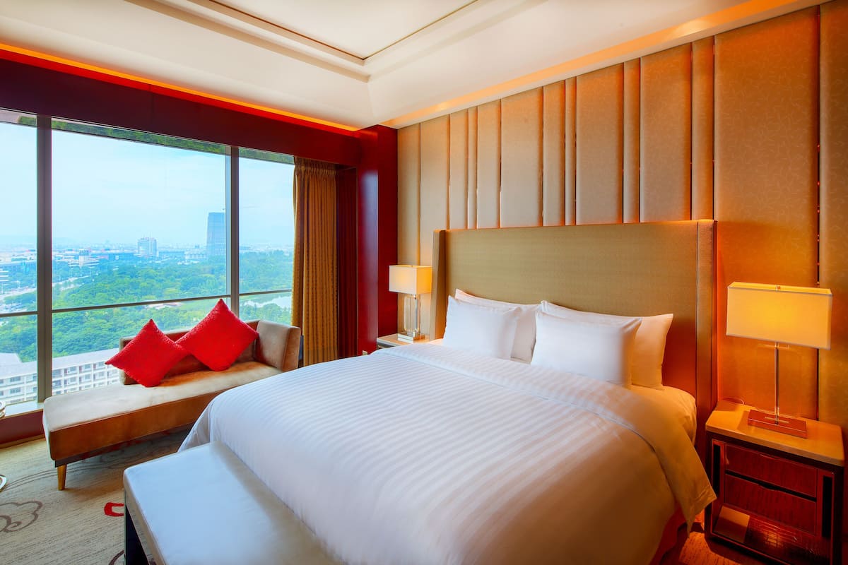 Ein großes Bett mit weißer Bettwäsche, ein Nachttisch mit Lampe, eine Bank mit roten Kissen und ein großes Fenster mit Blick auf die Stadt.