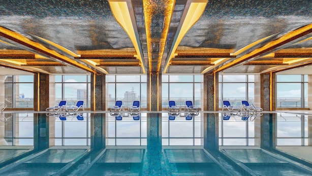 A large indoor swimming pool with clear blue water, surrounded by lounge chairs and large windows offering views of the city skyline. - Sanding New Century Grand Hotel Yiwu (Jinhua)