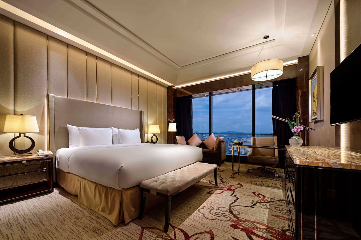 Ein Hotelzimmer mit einem großen Bett, einem Schreibtisch, einem Stuhl, einem kleinen Tisch mit Blumenvase und Blick auf die Stadt durch große Fenster.