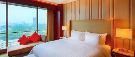 A hotel room with a large bed, a window with a city view, and a seating area with red cushions.