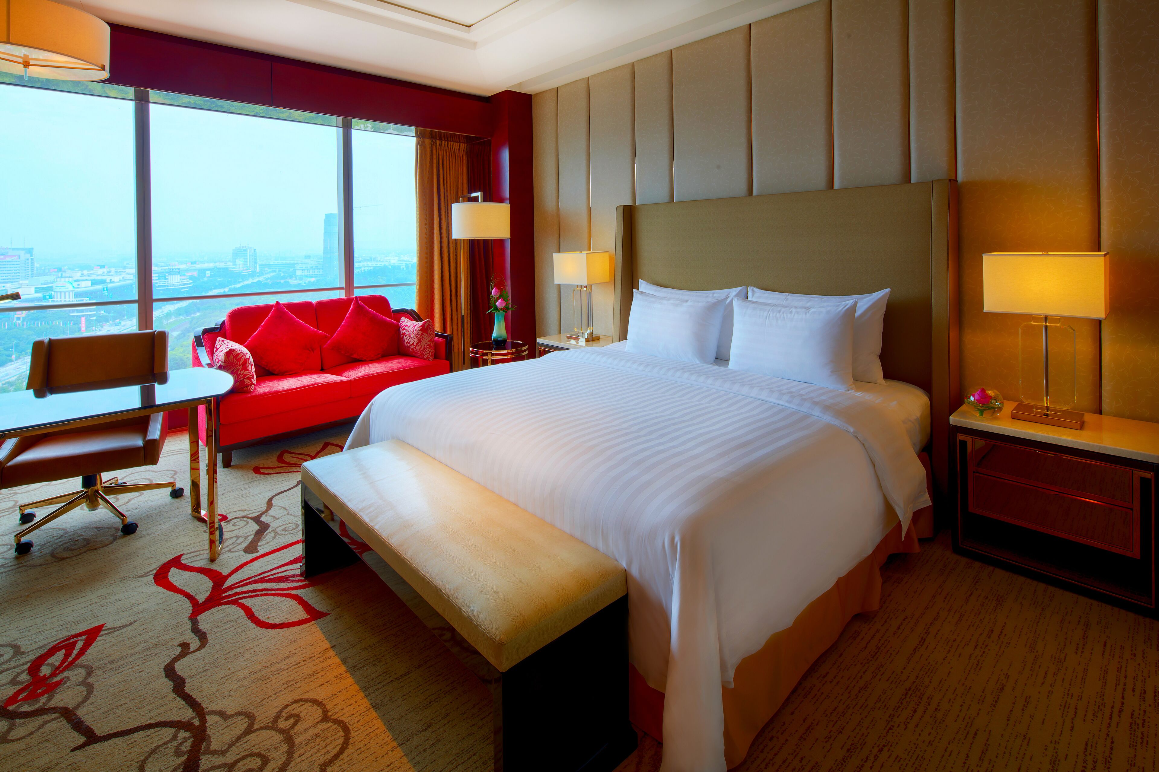 A hotel room with a large bed, a desk, a red sofa, and a view of the city.