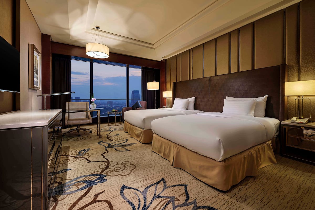 Ein Hotelzimmer mit zwei Betten, einem Schreibtisch, einem Stuhl, einem Fernseher und einem großen Fenster mit Blick auf die Stadt.