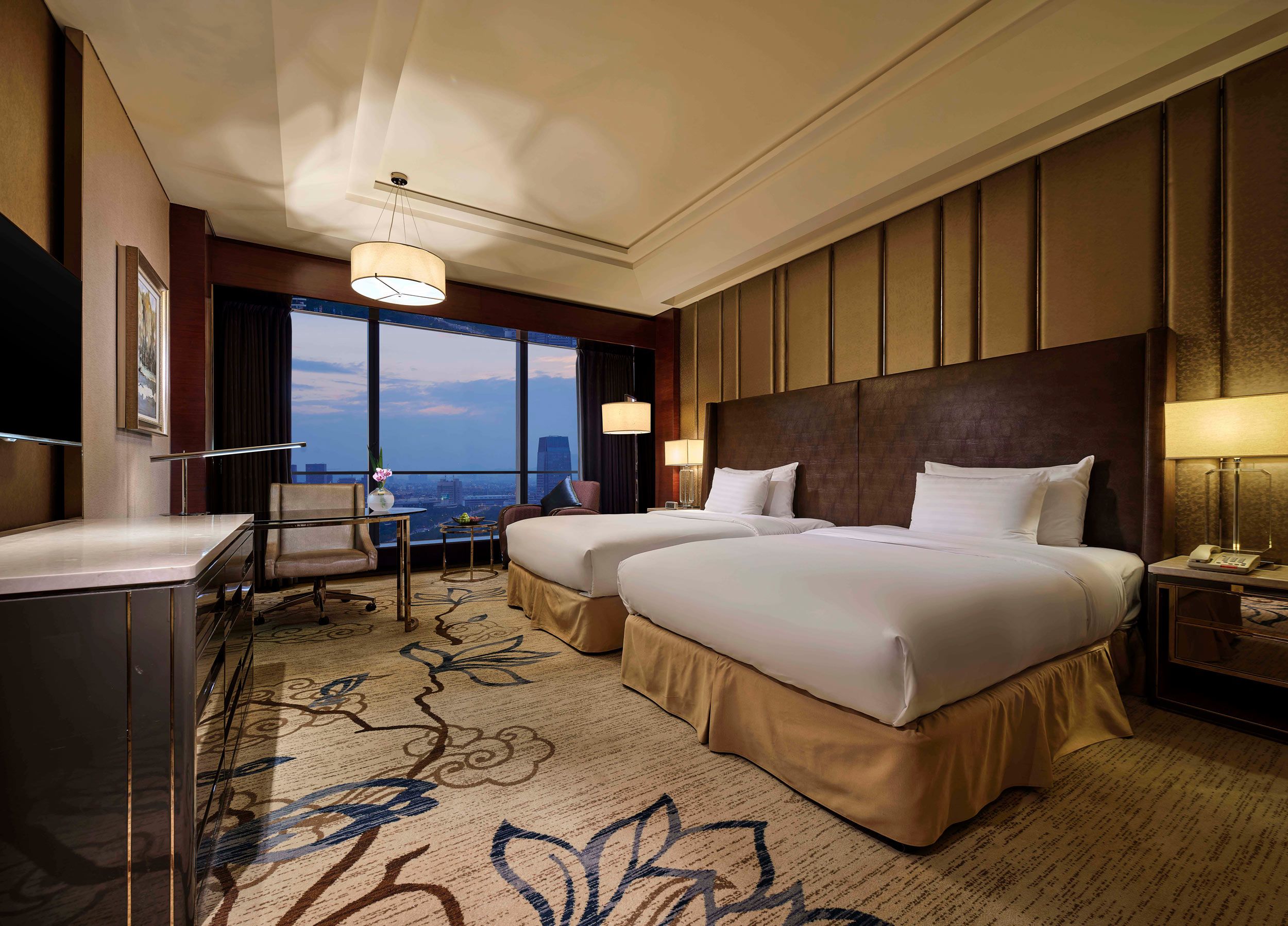 A hotel room with two beds, a desk, a chair, a TV, and a large window with a city view.