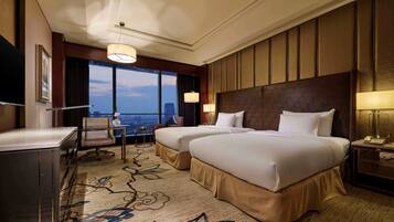 A hotel room with two beds, a desk, a chair, a TV, and a large window with a city view.