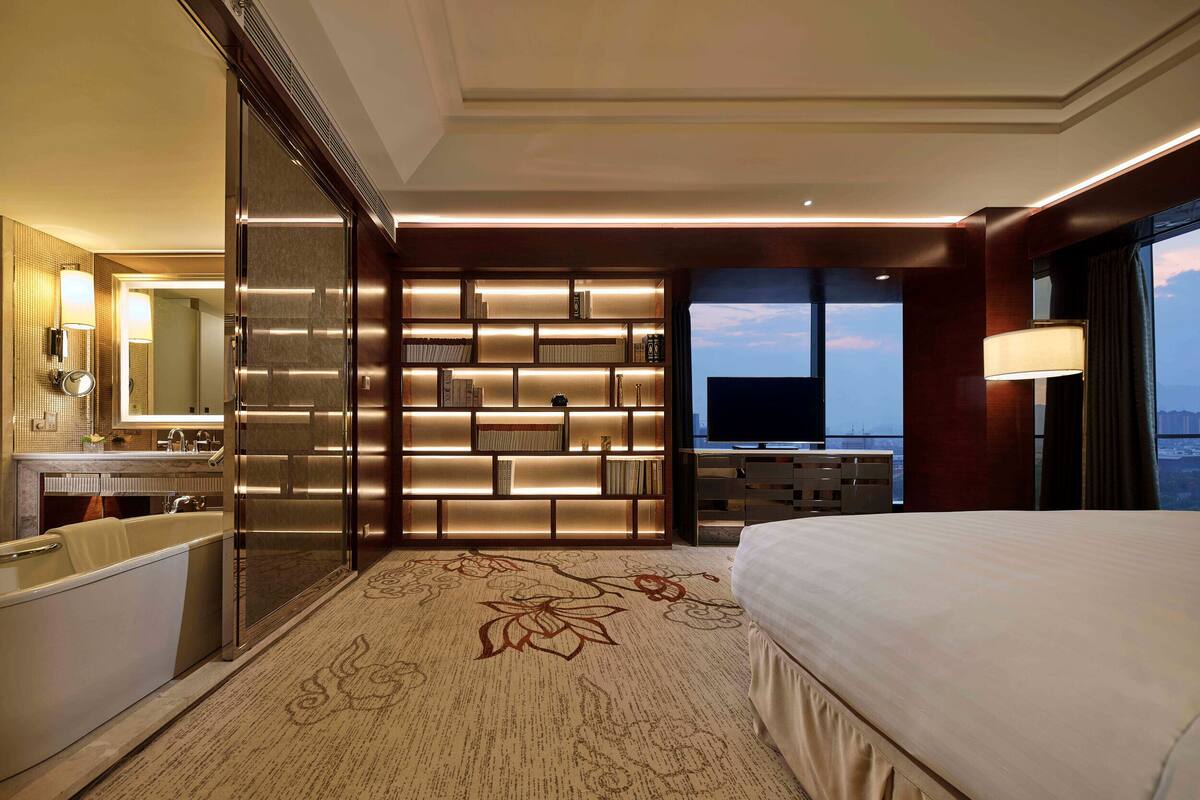 Ein modernes Hotelzimmer mit einem großen Bett, einem Schminktisch mit Spiegel, einem Bücherregal und einem Fernseher.