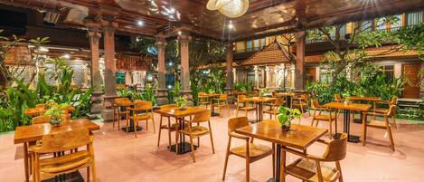 A spacious indoor area with wooden tables and chairs, surrounded by lush greenery and traditional architecture.