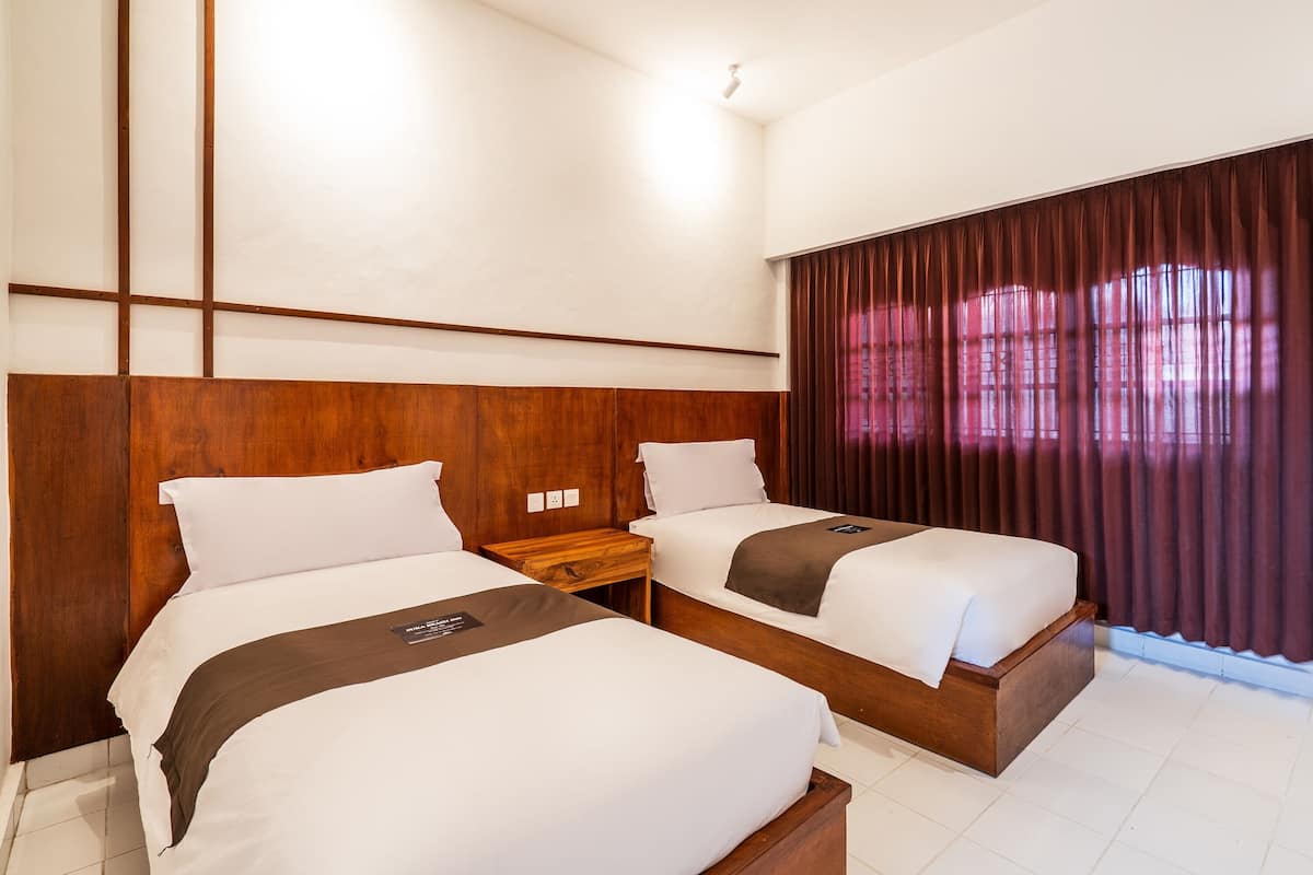 Habitación de hotel con dos camas, cabeceros de madera, una mesita de noche y un ventanal con cortinas.