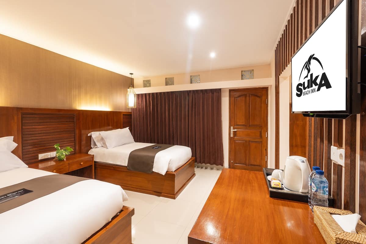 Habitación de hotel con dos camas, cabecero de madera, televisor de pantalla plana, mesita de noche con lámpara y bandeja con tetera y botella de agua.