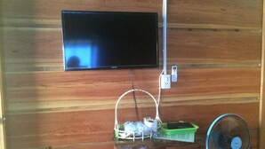 Un televisor montado en una pared de madera, un enchufe, una pequeña mesa con una cesta y un ventilador de pie.