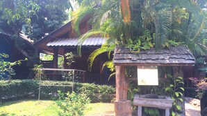 Una casa de madera con techo de tejas, rodeada de abundante vegetación y una pequeña estructura de madera con un letrero.