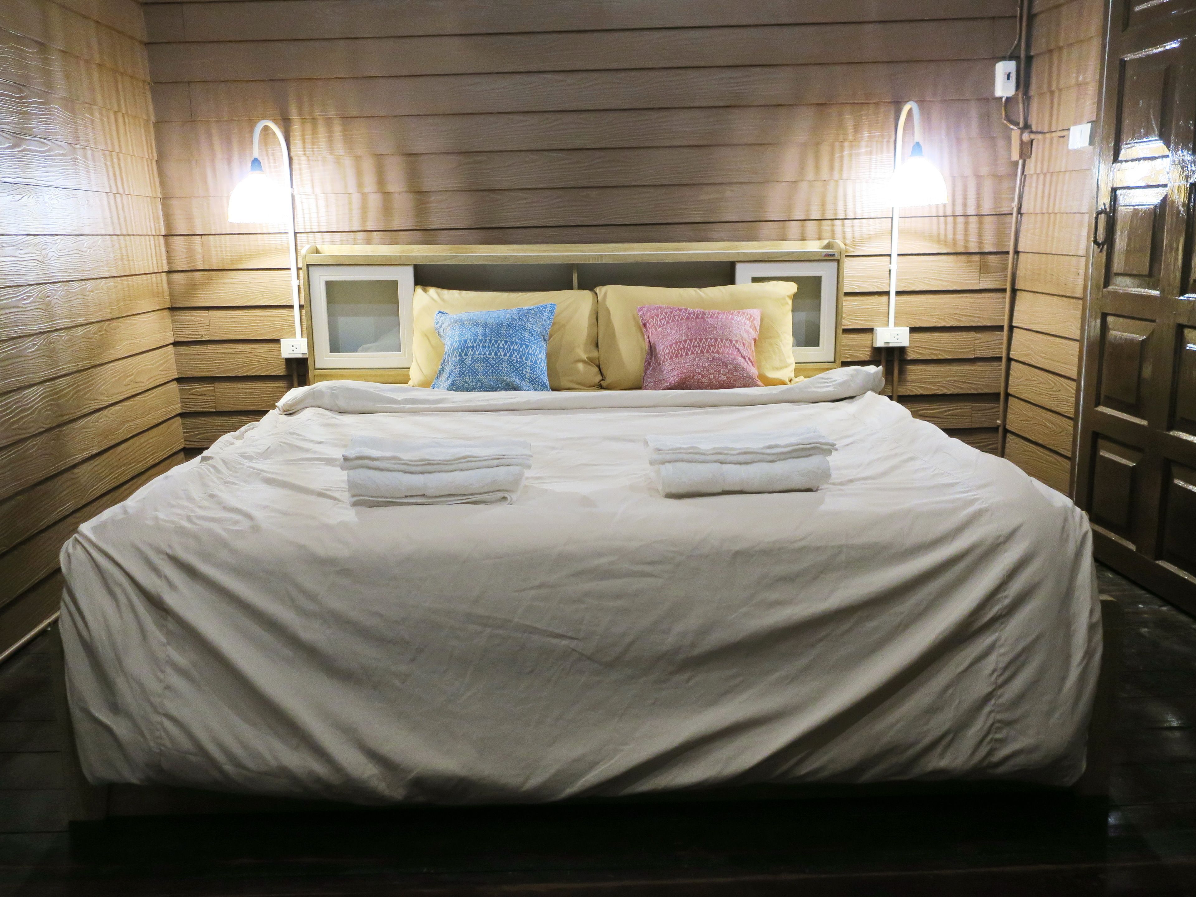 A neatly made bed with a white comforter and two folded towels, a wooden headboard, and two bedside lamps.