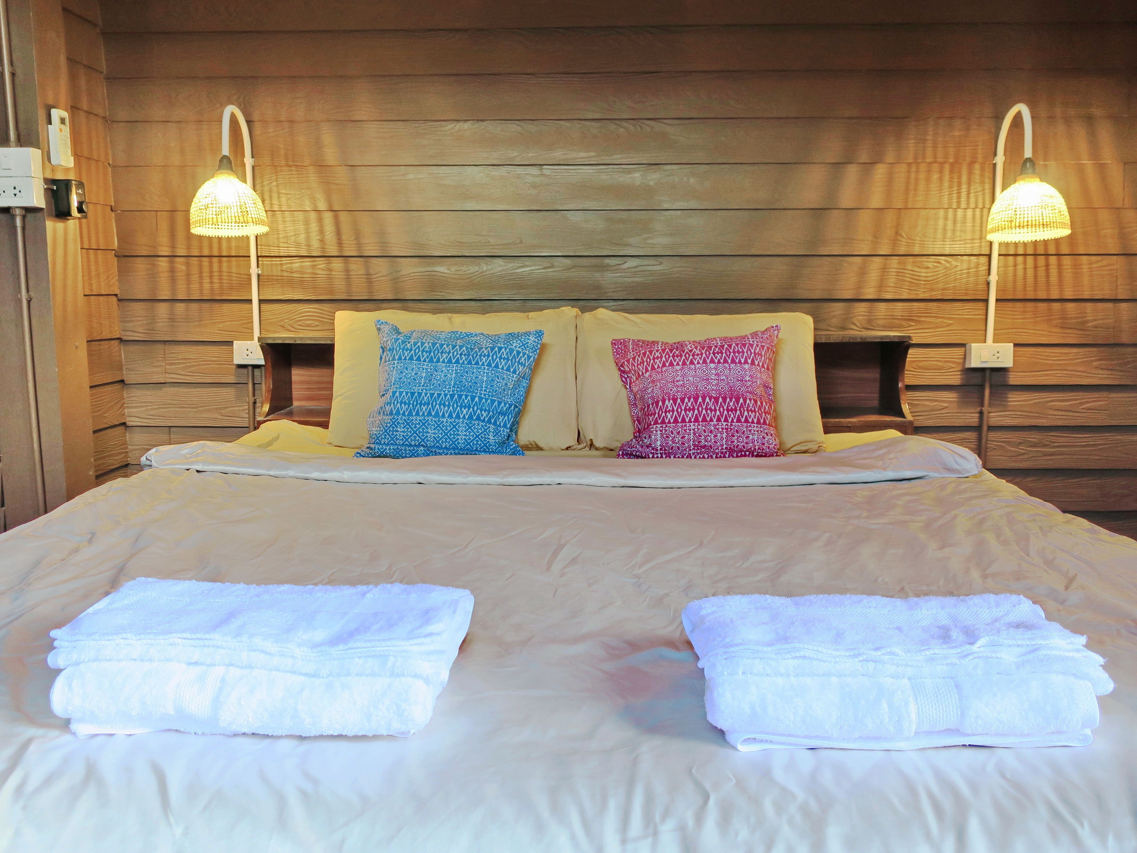 A neatly made bed with two pillows, two folded towels, and two wall-mounted lamps.