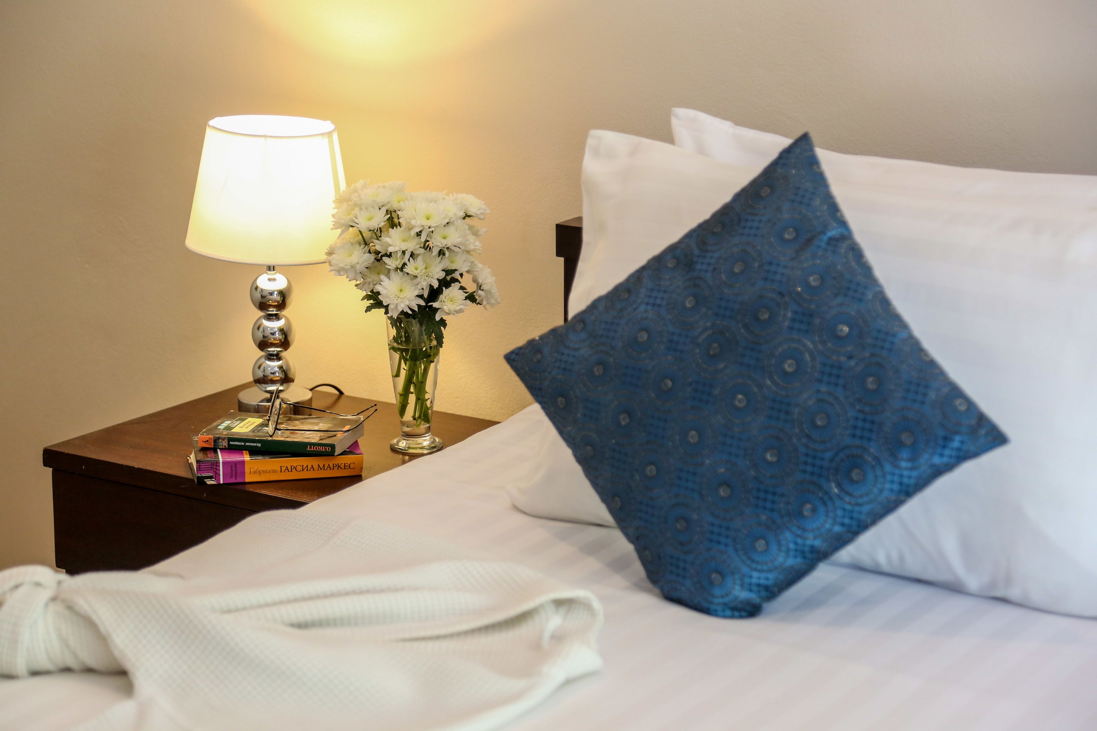 Una cama bien hecha con sábanas blancas, una almohada azul estampada, una lámpara de noche, un jarrón con flores blancas y una pila de libros sobre una mesita de noche de madera.
