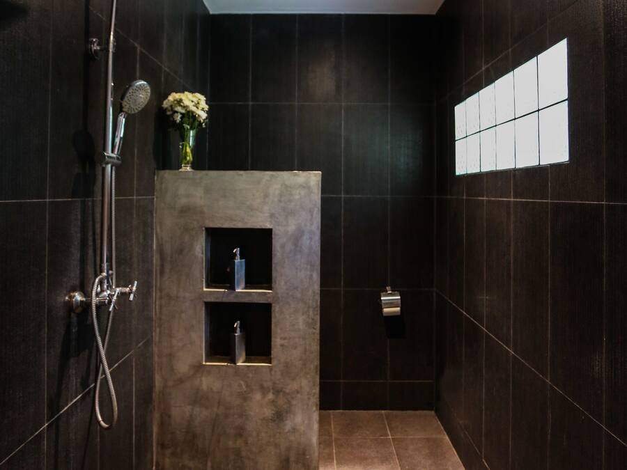 Un baño moderno con azulejos oscuros, una ducha con cabezal de ducha de mano, un estante pequeño con artículos de aseo y una ventana con diseño de cuadrícula.