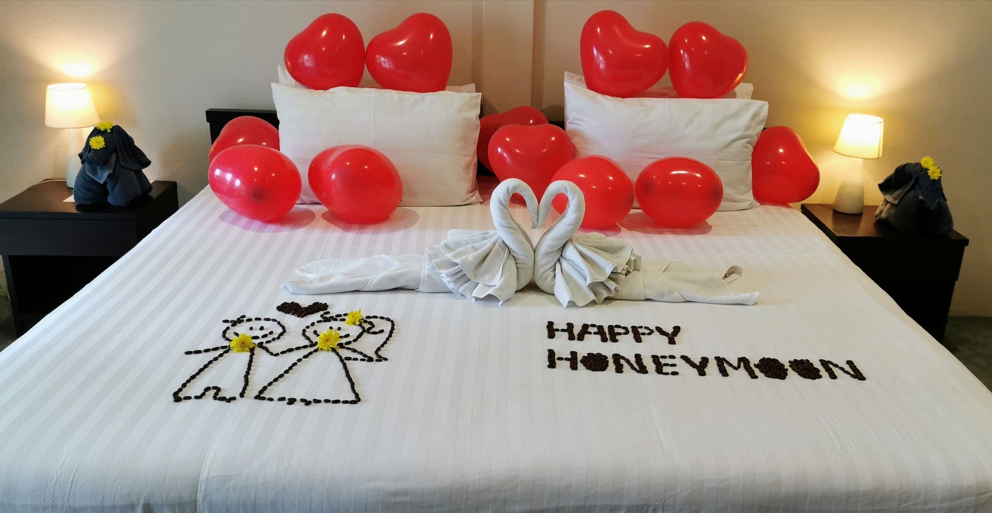 A hotel room with a bed adorned with red heart-shaped balloons, a bouquet of flowers, and a 'Happy Honeymoon' message.