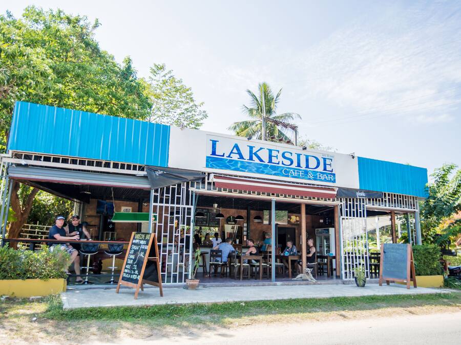 Un café y bar junto al lago con mesas al aire libre y un toldo azul.