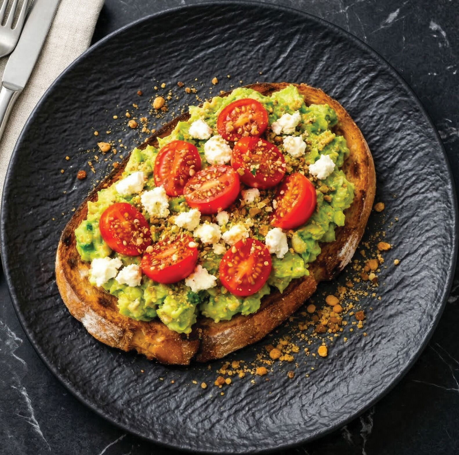Una tostada con aguacate, tomates y queso sobre un plato oscuro.