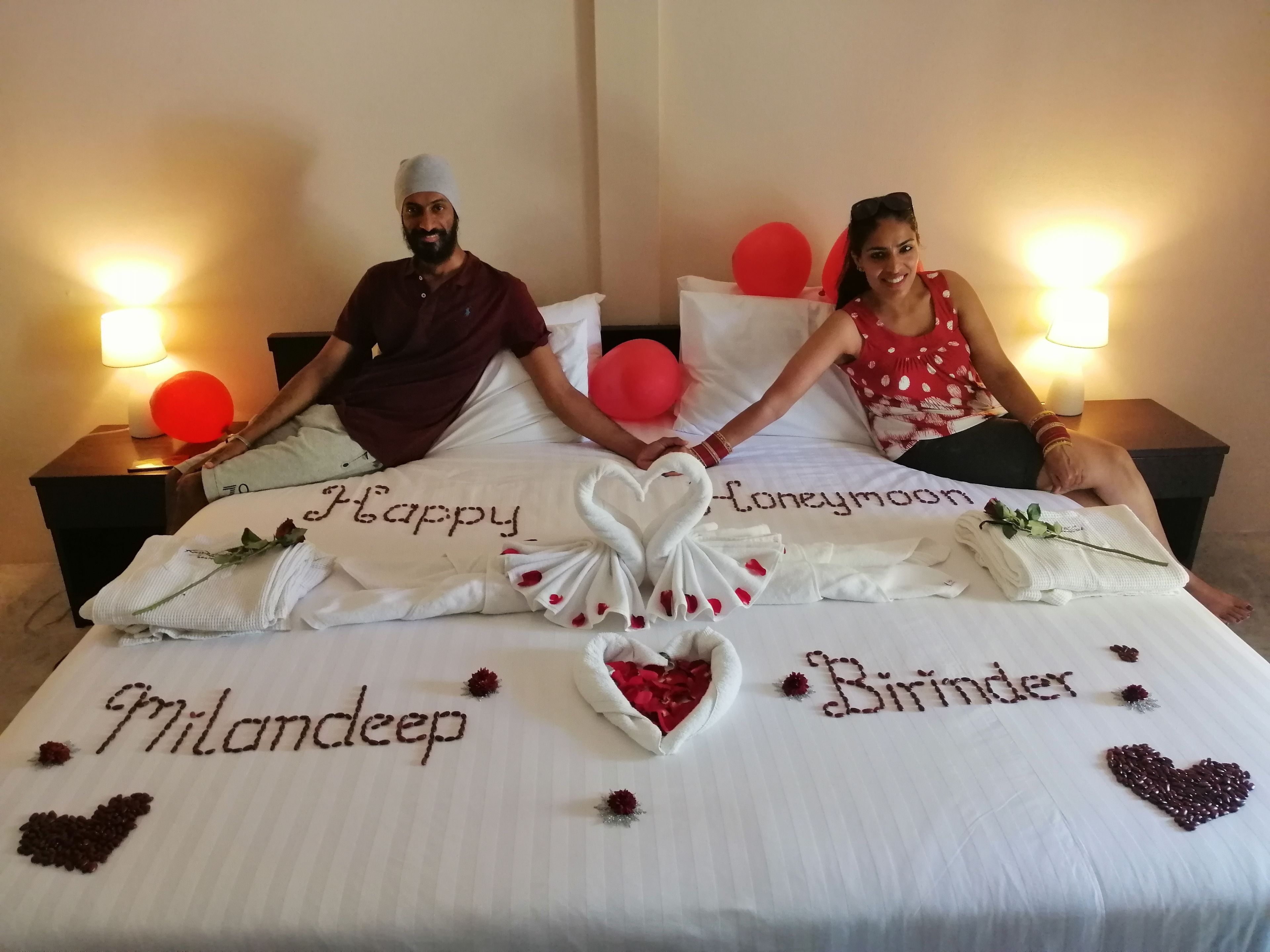 A bed with personalized decorations including the names "Milandeep" and "Birinder" and the words "Happy Honeymoon" and "Birthday".