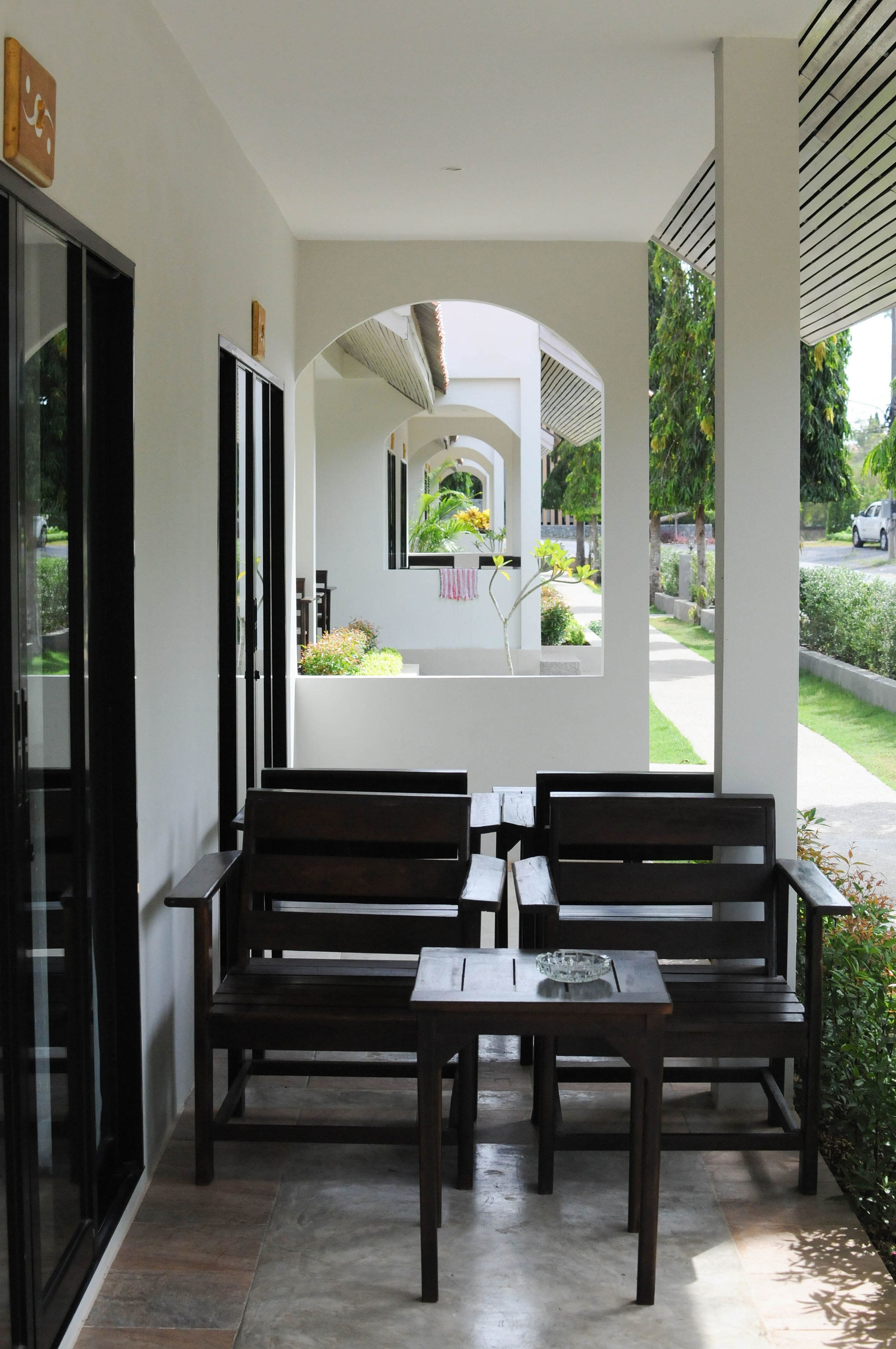 A covered outdoor seating area with benches and a table, a glass door, and a view of a garden and street.