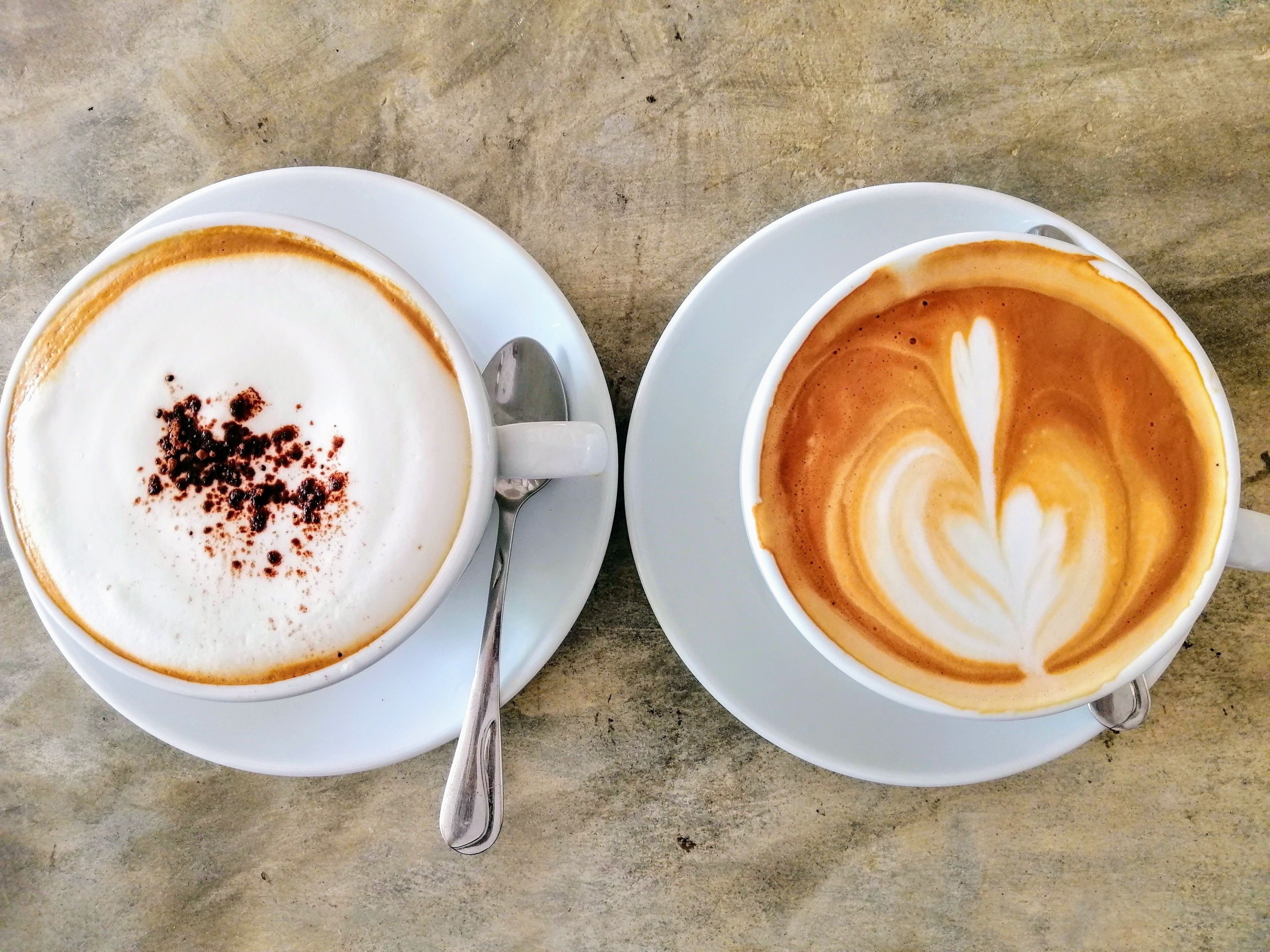 Duas chávenas de café sobre pires brancos, com colheres, pousadas sobre uma superfície texturizada.