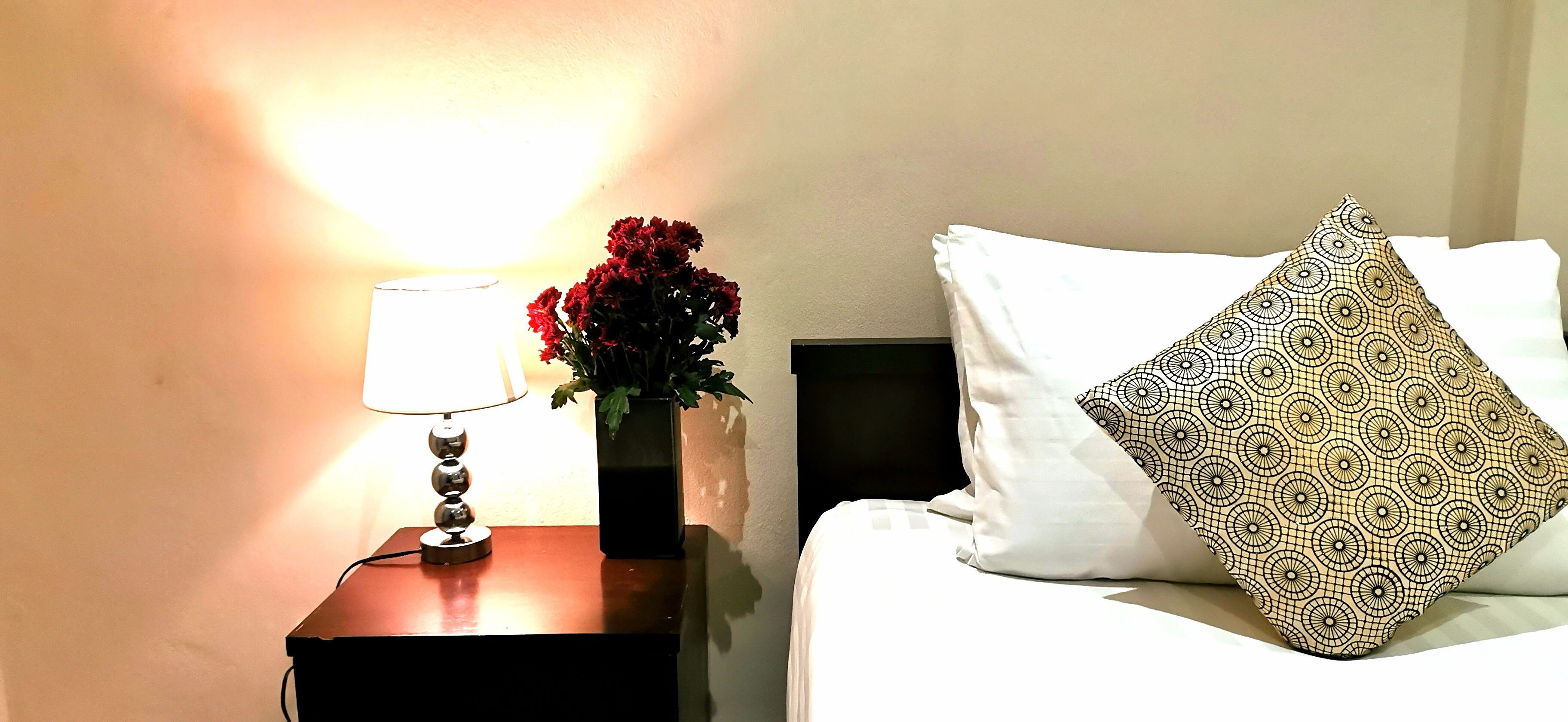 Una cama bien hecha con una almohada estampada y un florero con flores rojas sobre una mesita de noche junto a una lámpara.
